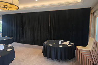 Black event draping across a banquet room wall with round dining tables, black tablecloths and neutral carpet flooring