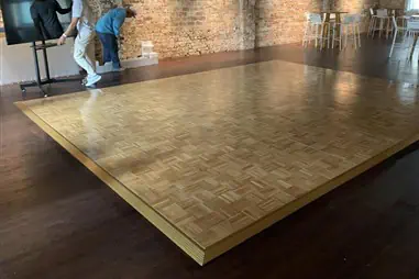 A parquet dance floor with geometric wood pattern ready for hire, positioned in an industrial warehouse space with exposed brick walls and high table seating