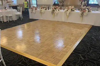 A wooden parquet dance floor with herringbone pattern positioned in front of an elegant white bridal table decorated with cascading floral arrangements in soft pink and cream tones, set in a reception venue with white chiavari chairs and large windows