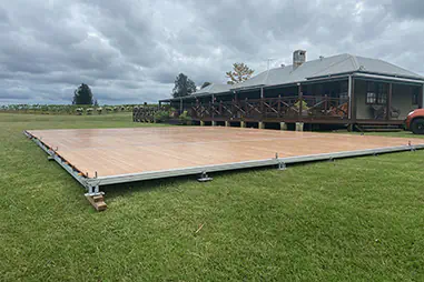 Large outdoor event flooring platform installed on grass beside a rural venue, with timber-look panels, metal edging and surrounding vineyard landscape