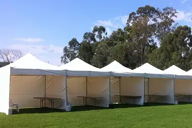 Row of white pop-up canopies set up on grass with side walls and open fronts, creating a covered outdoor event area beside trees