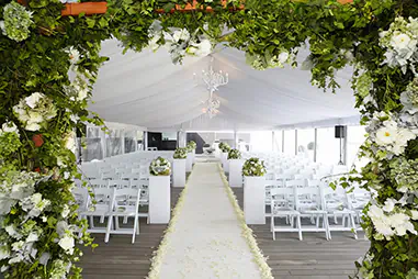 Wedding marquee ceremony setup with white guest seating, a central aisle runner, floral arch entrance, white draped ceiling, chandelier lighting and greenery with white flowers framed around the doorway