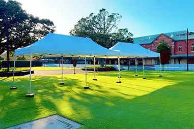 White outdoor marquees set up on a green lawn beside a red brick venue, providing open covered shelter for an outdoor event area