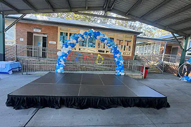 Covered outdoor stage with black skirting positioned beneath a blue and white balloon arch in a school event area