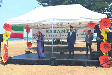 Festival stage cover with a white canopy, black skirted platform, speakers, microphones and decorative flowers for a coastal outdoor community event