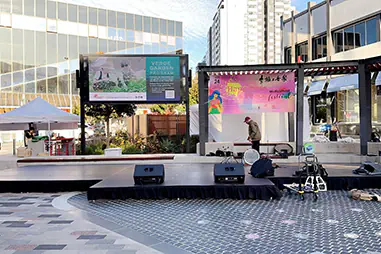 Outdoor event stage setup in a city plaza with modular black platforms, audio speakers and festival signage in the background