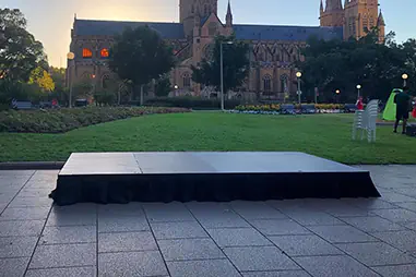 Outdoor stage platform set on a paved area beside a lawn, with a large cathedral building in the background at dusk