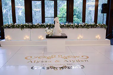 Wedding stage with a white bridal table, soft floral styling, candles, tiered cake display and a glossy white dance floor with custom couple lettering